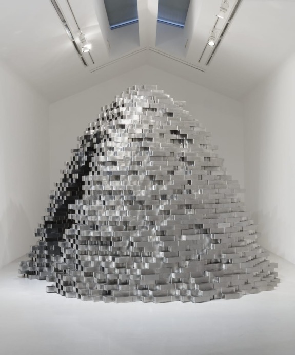 Large installation of stacked metallic blocks forming a mound in a white gallery space with overhead lighting.