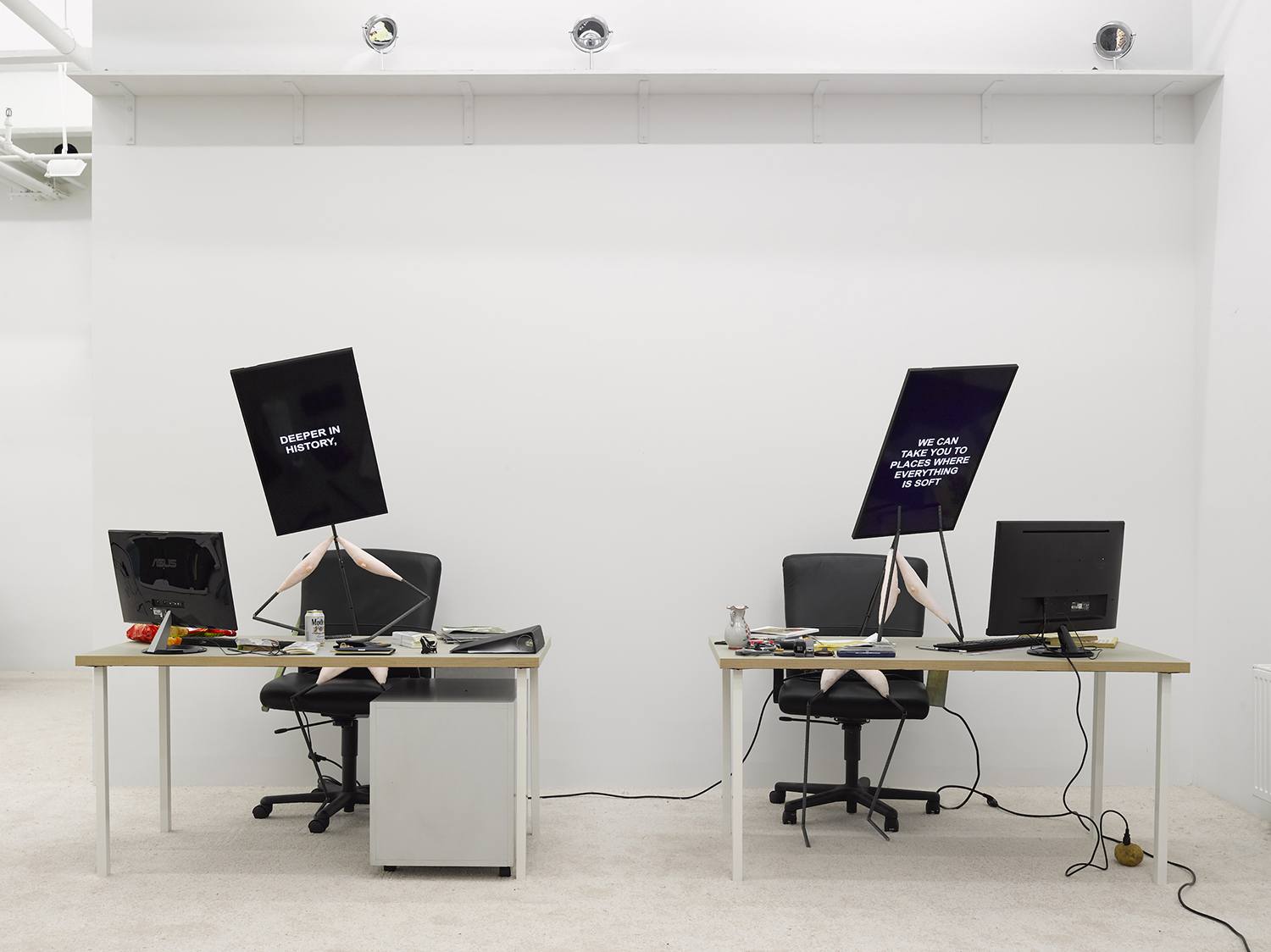 Two office desks with computer monitors held up by mannequin arms in a white room, displaying text messages on the screens.