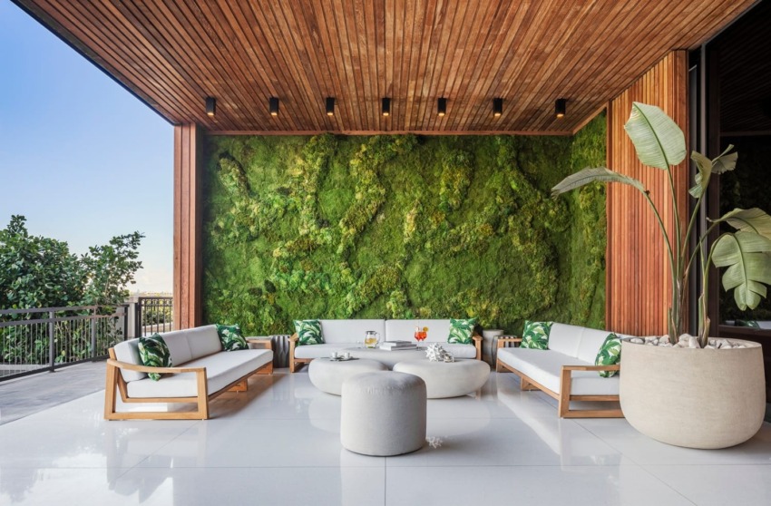Modern outdoor patio with wooden ceiling, green moss wall, white seating, and potted plants.