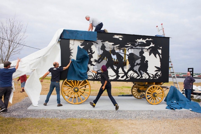 People installing a white sheet on a vintage wagon with cutout silhouette art, revealing a scene of figures and foliage.