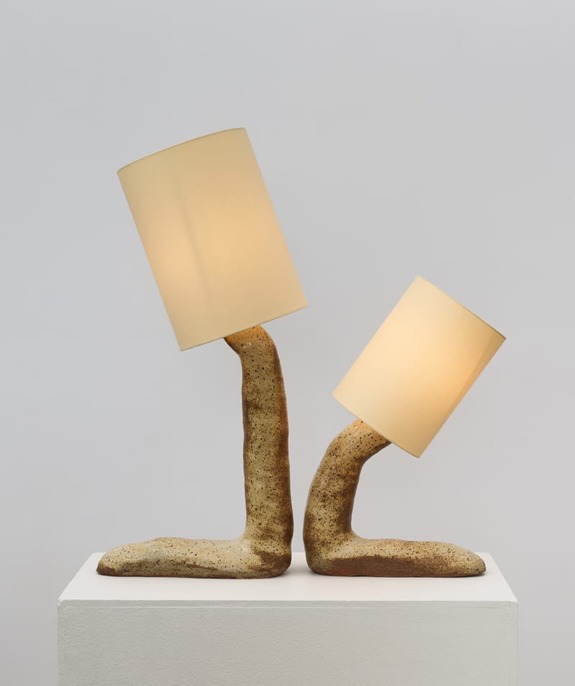 Two beige, sculptural lamps on a plinth with organic, curved bases and cylindrical shades illuminating softly.