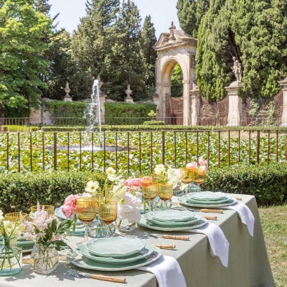 Elegant outdoor dining setup with decorative tableware in a garden setting, featuring a fountain and historical archway.