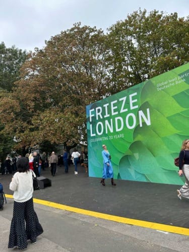 People walking near a large green Frieze London 20th anniversary sign outdoors with trees in the background.