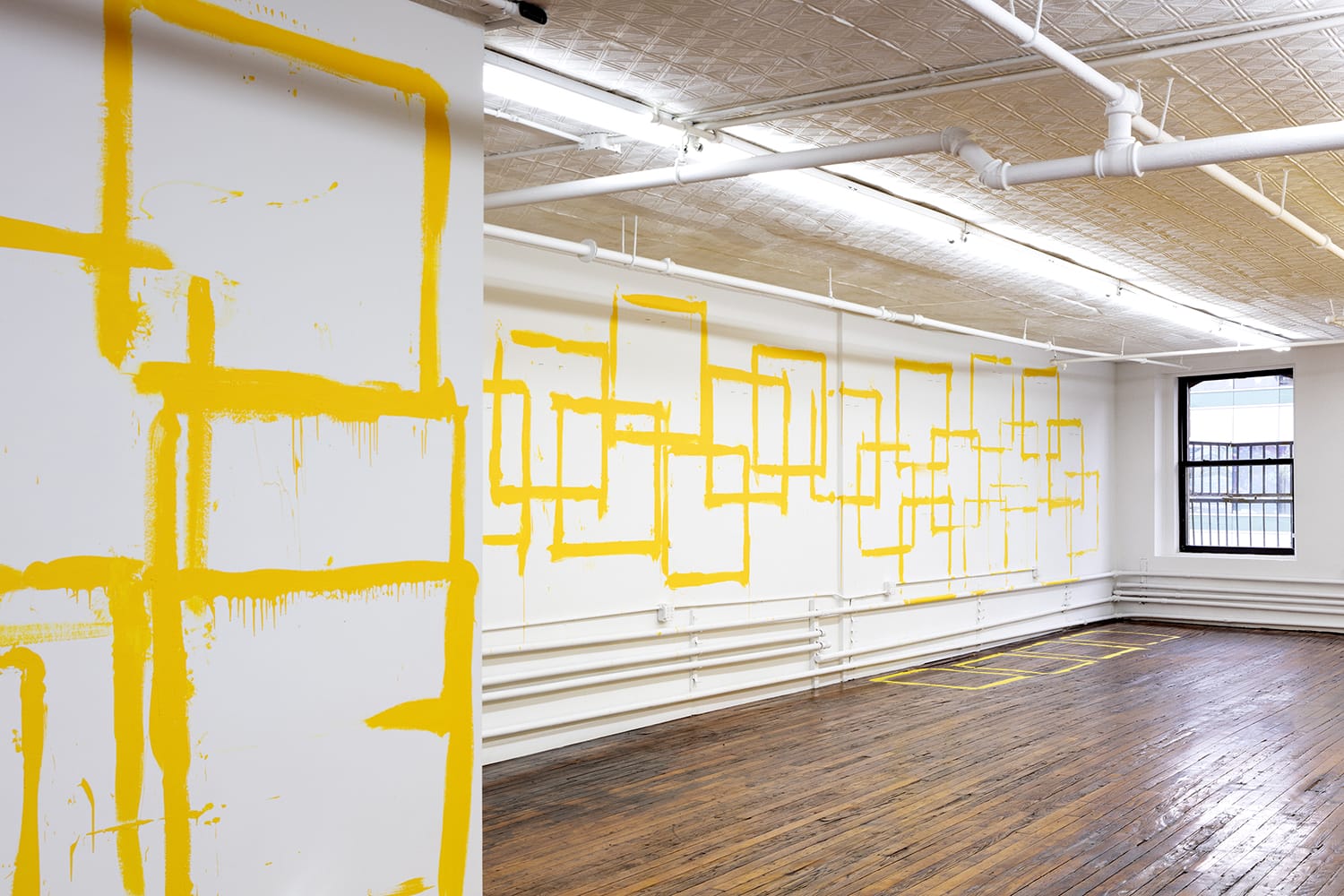 Art installation with bold yellow geometric shapes painted on white walls of an industrial-style room with wooden floors.