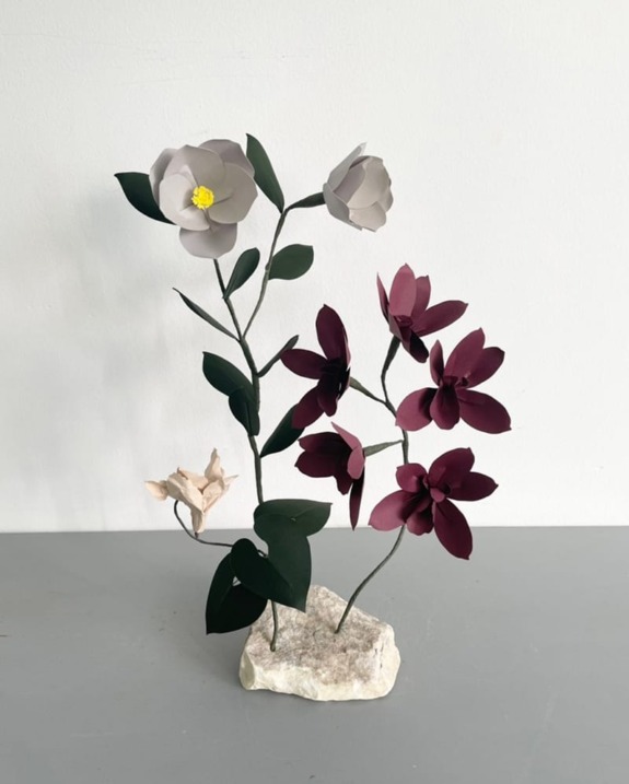 Paper flowers in white and burgundy with green leaves arranged on a stone base against a plain background.