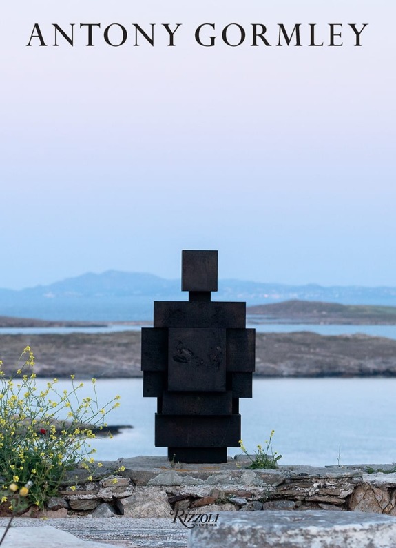 Statue of geometric figure by water with mountains in background under Antony Gormley text.