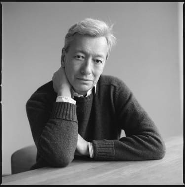 Black and white portrait of a person with short hair sitting at a table, resting their head on their hand, looking at the camera.