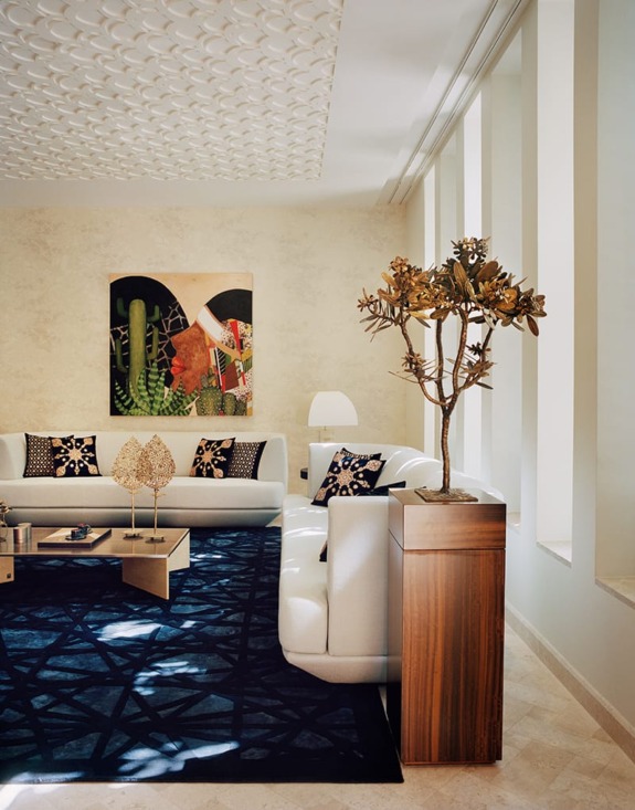 Modern living room with white sofas, decorative cushions, a geometric rug, abstract wall art, and a tall plant on a wooden stand.