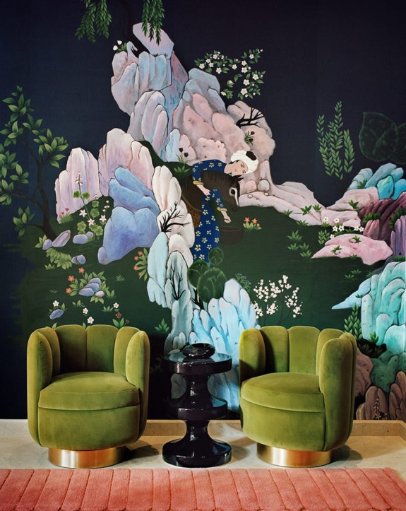 Two green velvet chairs and a black side table in front of a colorful, artistic mural with nature and figure elements.