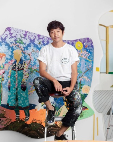 Artist sitting on a stool in front of colorful artwork, wearing a paint-splattered outfit.