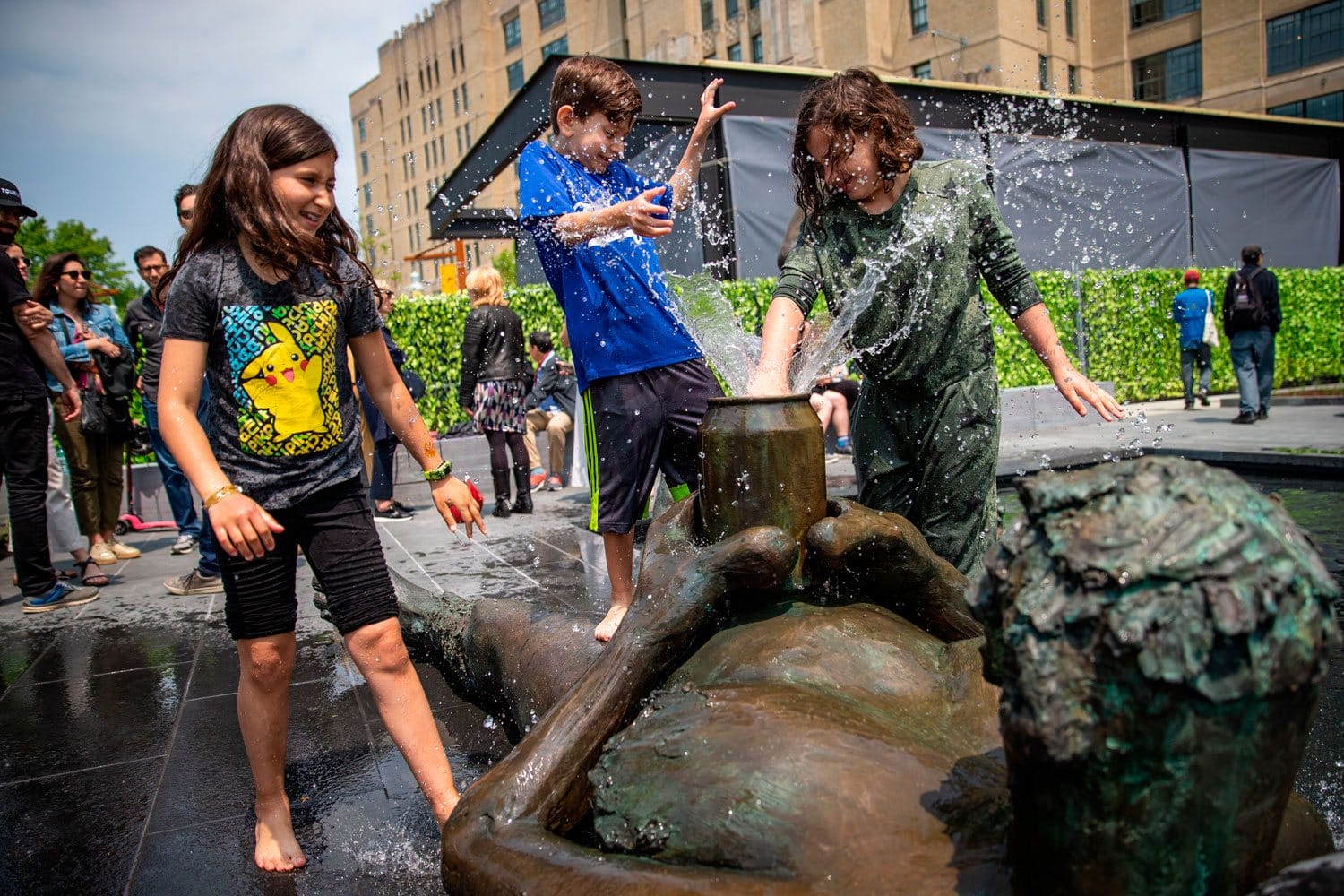 Children play in a fountain with a large sculpture, laughing and splashing water, surrounded by a crowd in a city setting.