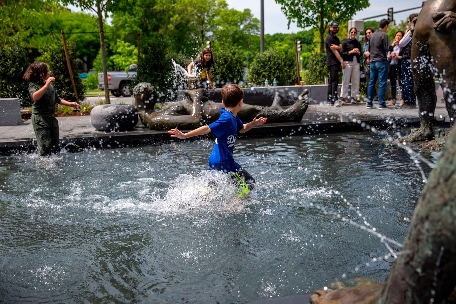 Child in a blue shirt splashing water in a fountain with people watching in the background on a sunny day.