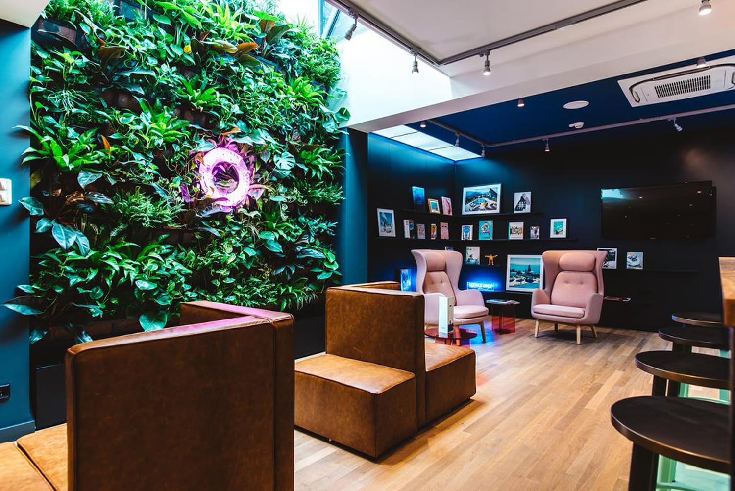 Modern office lounge with vertical garden, brown seating, artworks on dark walls, and a skylight for natural light.