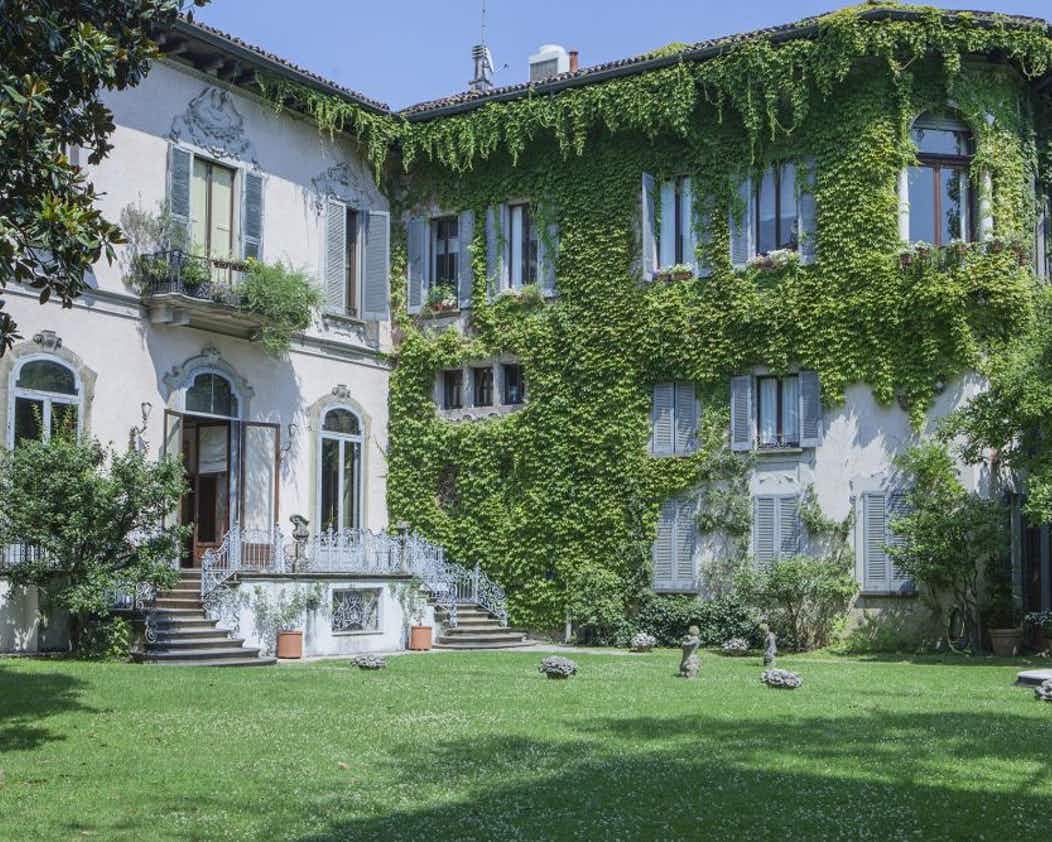 Large ivy-covered villa with white shutters, ornate balcony, and lush green garden in sunny weather.