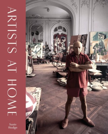 Artist standing in a studio filled with paintings and art supplies, surrounded by creativity and color.