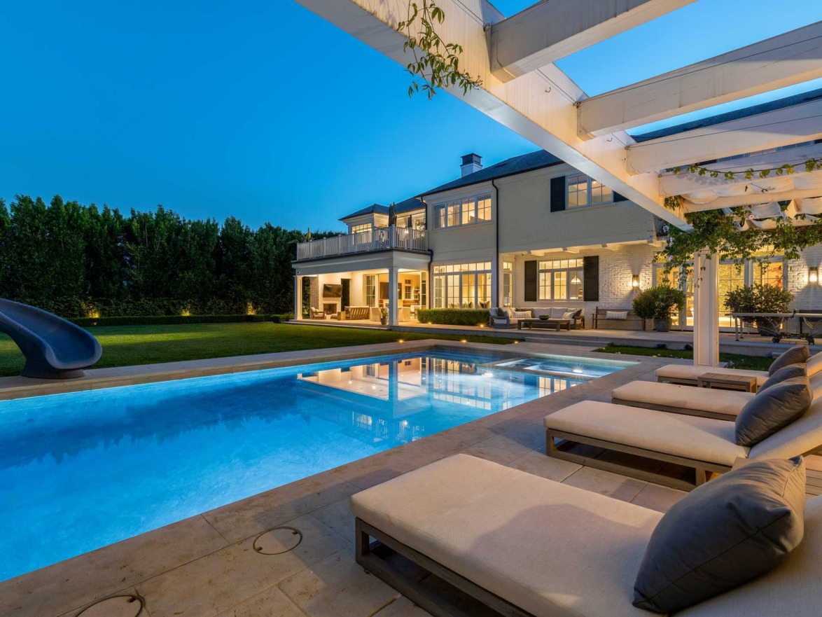 Luxurious backyard with illuminated swimming pool, lounge chairs, and modern house at dusk.