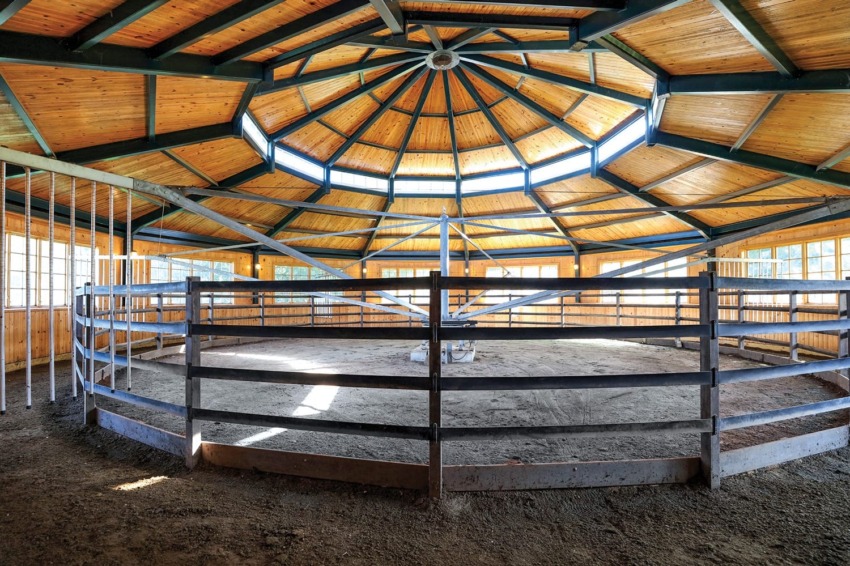 Interior of a wooden round pen for horse training with a high ceiling and support beams in a spacious, well-lit area.