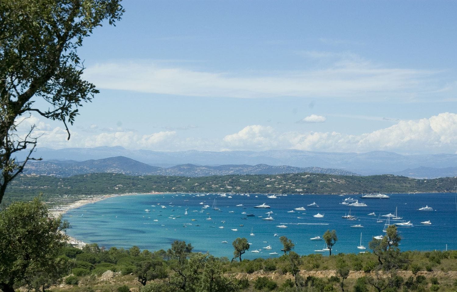 Boats anchored in a calm bay with lush green hills and distant mountains under a blue sky with scattered clouds.