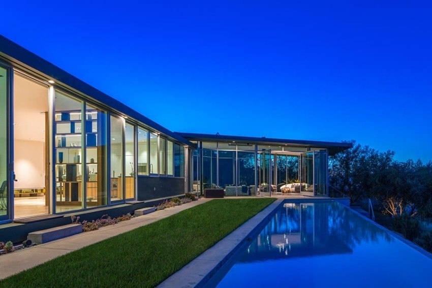Modern glass house with large windows next to a reflective swimming pool at dusk, surrounded by lush greenery.