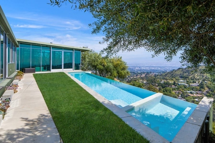 Modern house with glass windows overlooking a cityscape, featuring an infinity pool and surrounding greenery.