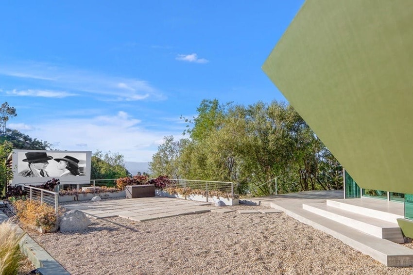 Modern outdoor patio with gravel, sleek green structures, and a large art piece against a backdrop of trees and blue sky.
