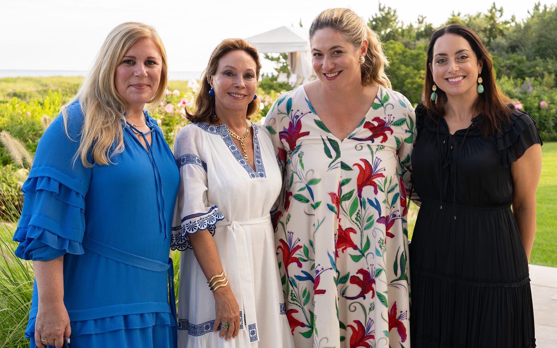 Four women smiling outdoors, standing together in colorful dresses, with greenery and a white tent in the background.