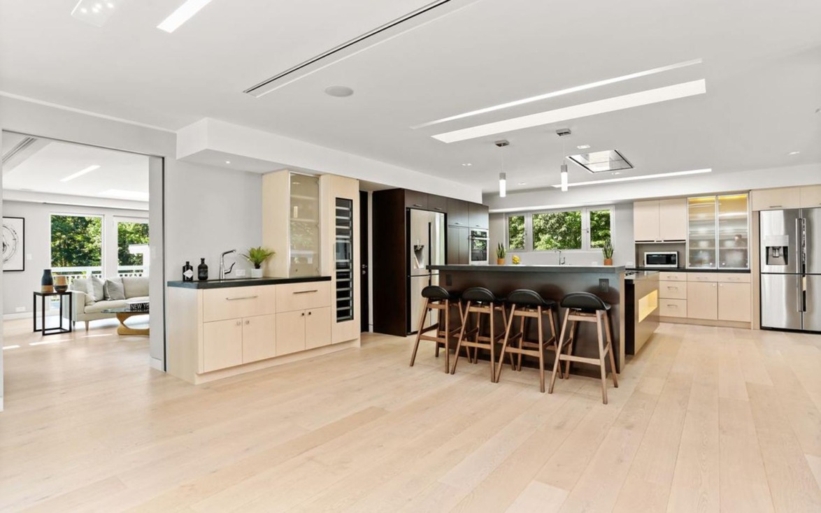 Modern kitchen with light wood flooring, large island, black stools, stainless steel appliances, and ample natural light.