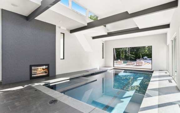 Indoor pool area with modern design, skylights, and a fireplace, overlooking an outdoor patio through large windows.
