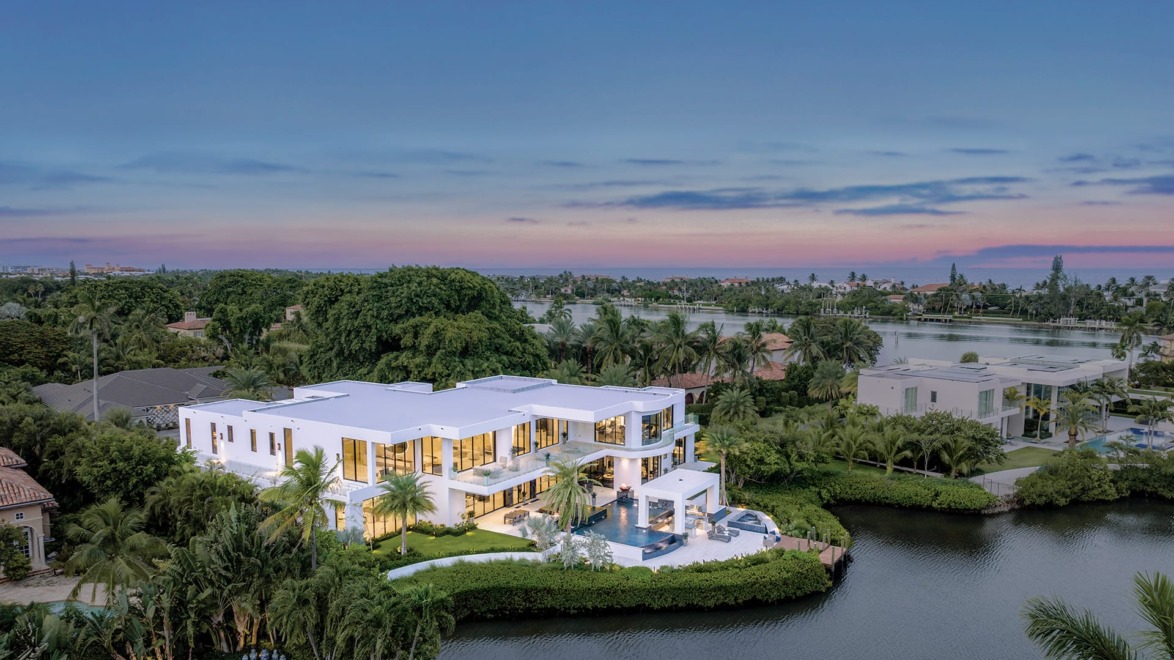 Aerial view of a modern white mansion by a waterway, surrounded by lush greenery and a serene landscape at sunset.