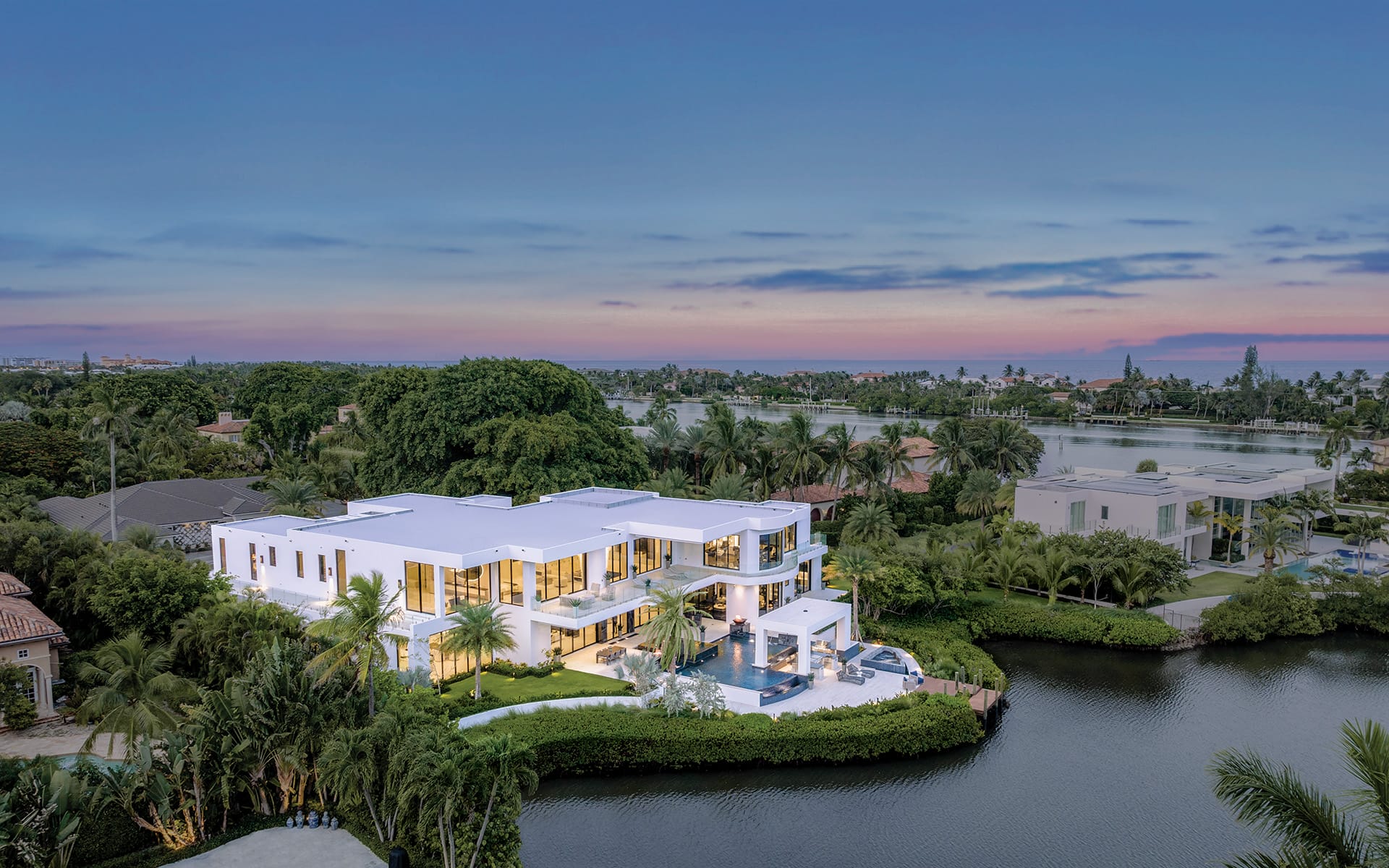 Aerial view of a modern white mansion by a waterway, surrounded by lush greenery and a serene landscape at sunset.