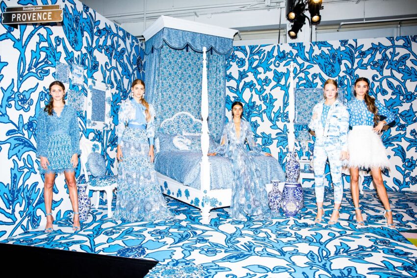 Models pose in a room decorated with blue and white floral patterns, including a canopy bed, rugs, and walls.