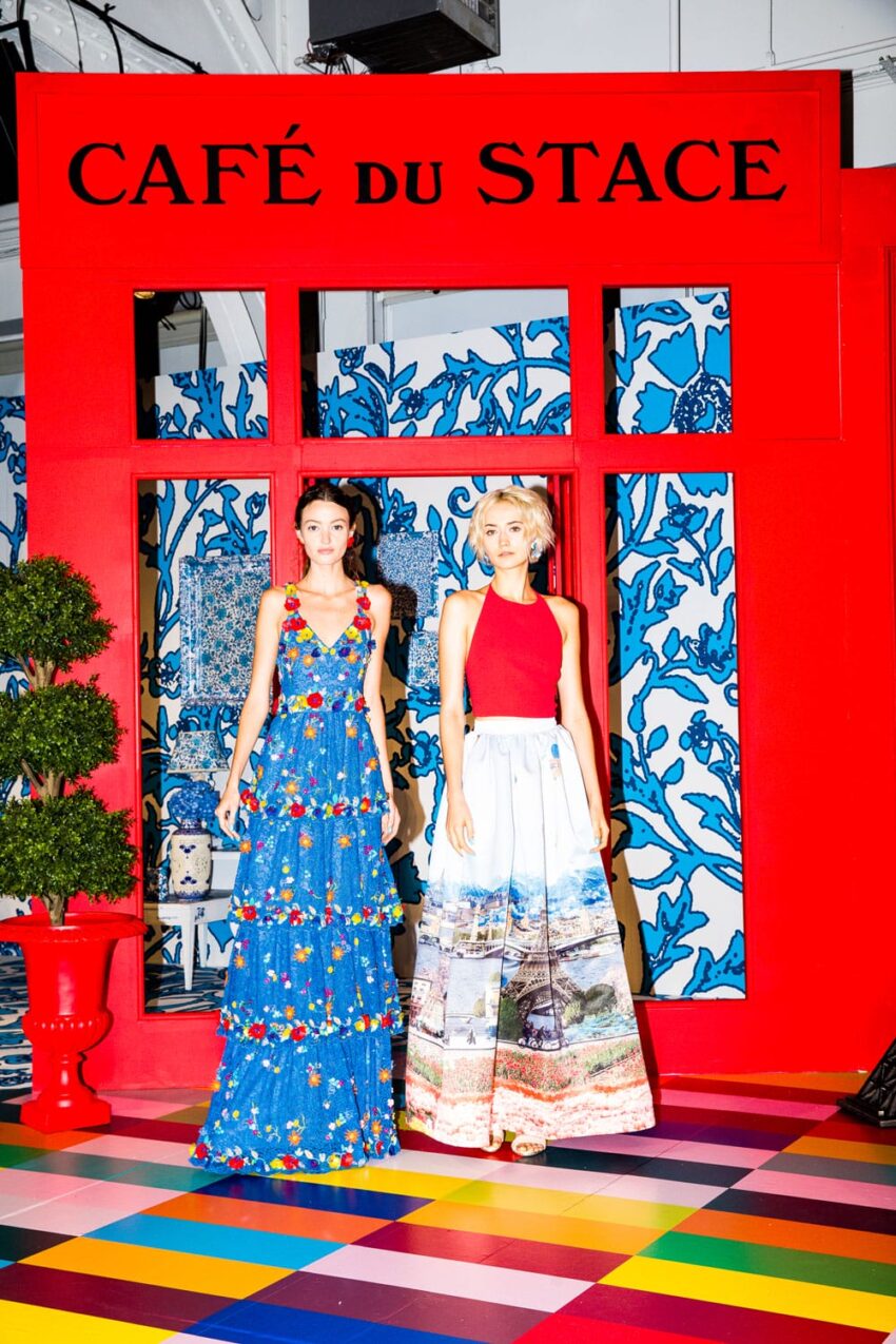 Two models in vibrant dresses standing in front of a colorful Café Du Stace backdrop with a patterned blue and white design.