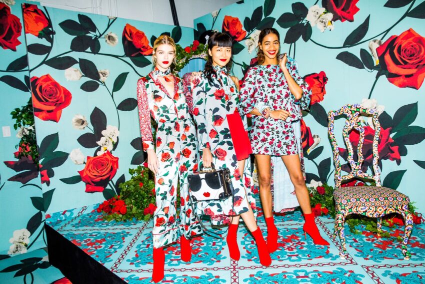 Three models in floral dresses and red boots pose in a vibrant room with floral patterns and decor.