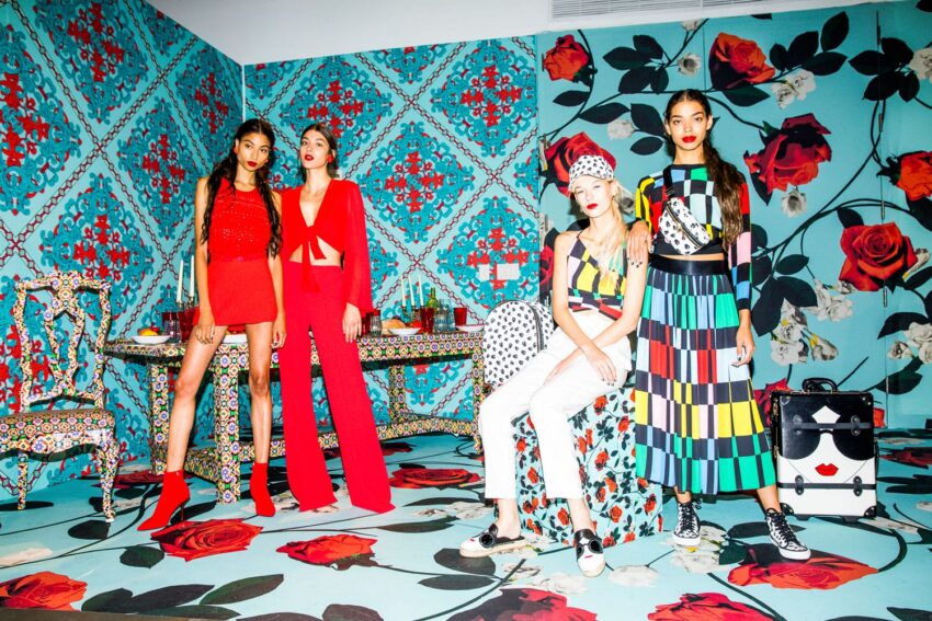 Four fashionable women in bold, colorful outfits posing in a room with vibrant floral wallpaper and decor.