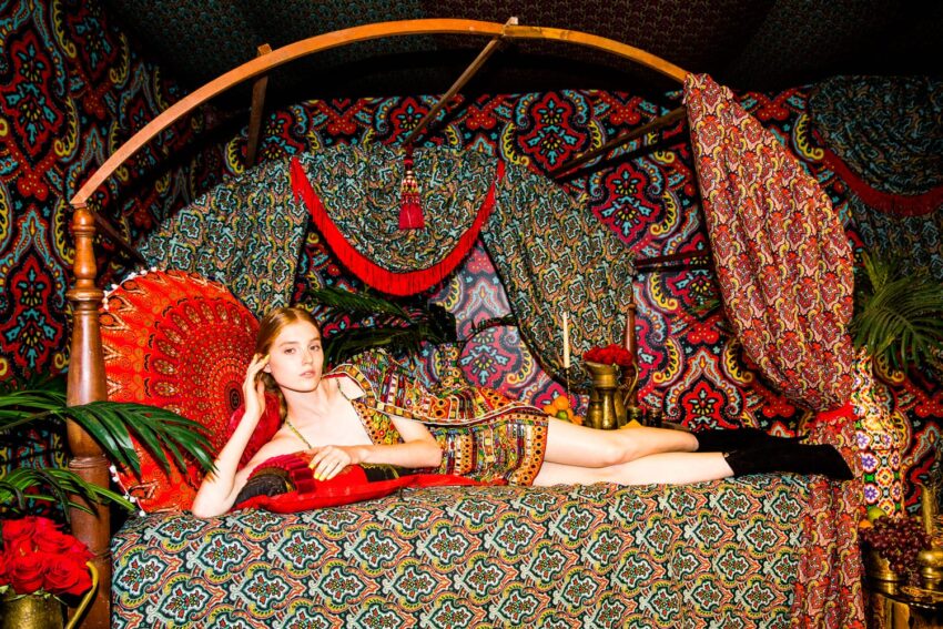 A woman in a patterned dress reclines on a bed surrounded by vibrant, colorful patterns and lush plants.