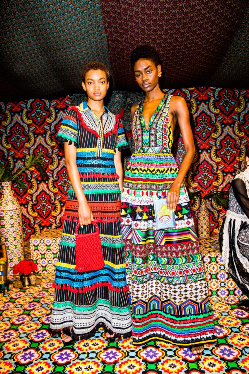Two models in colorful, patterned dresses standing on a vibrant, patterned floor with a patterned backdrop.