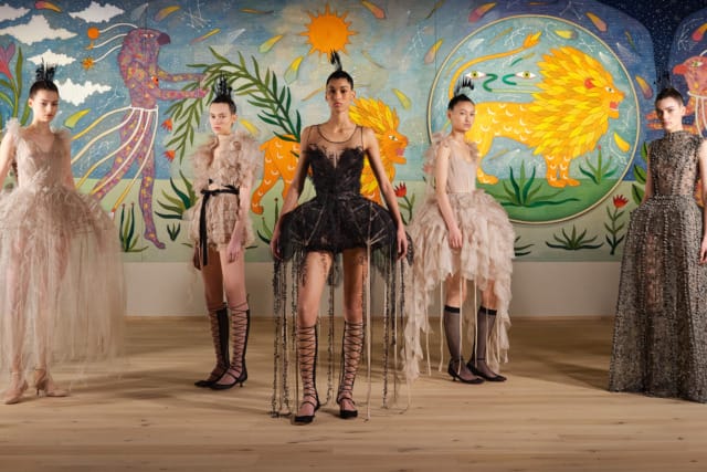 Fashion models in unique, artistic dresses stand in front of a colorful mural with birds and a lion.