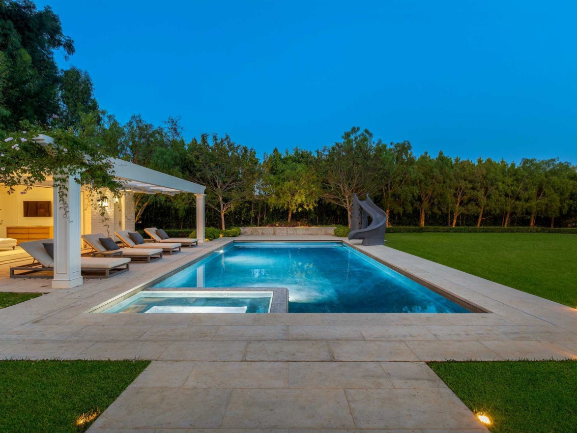Luxury outdoor pool with lounge chairs and water slide, surrounded by greenery at dusk.