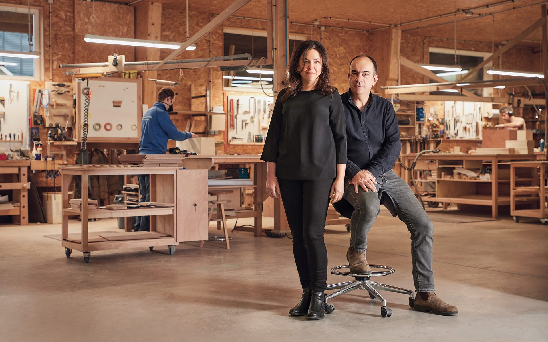 Two people in a woodworking workshop with tools and projects in the background.
