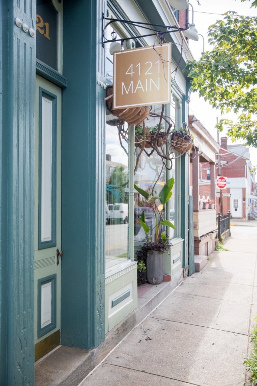 Street view of a quaint café entrance with a sign reading "4121 Main" and potted plants by the window.