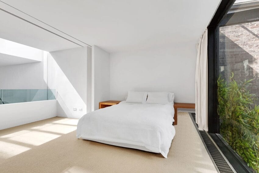 Minimalistic bedroom with a white bed, large window, and natural light streaming onto the carpeted floor.