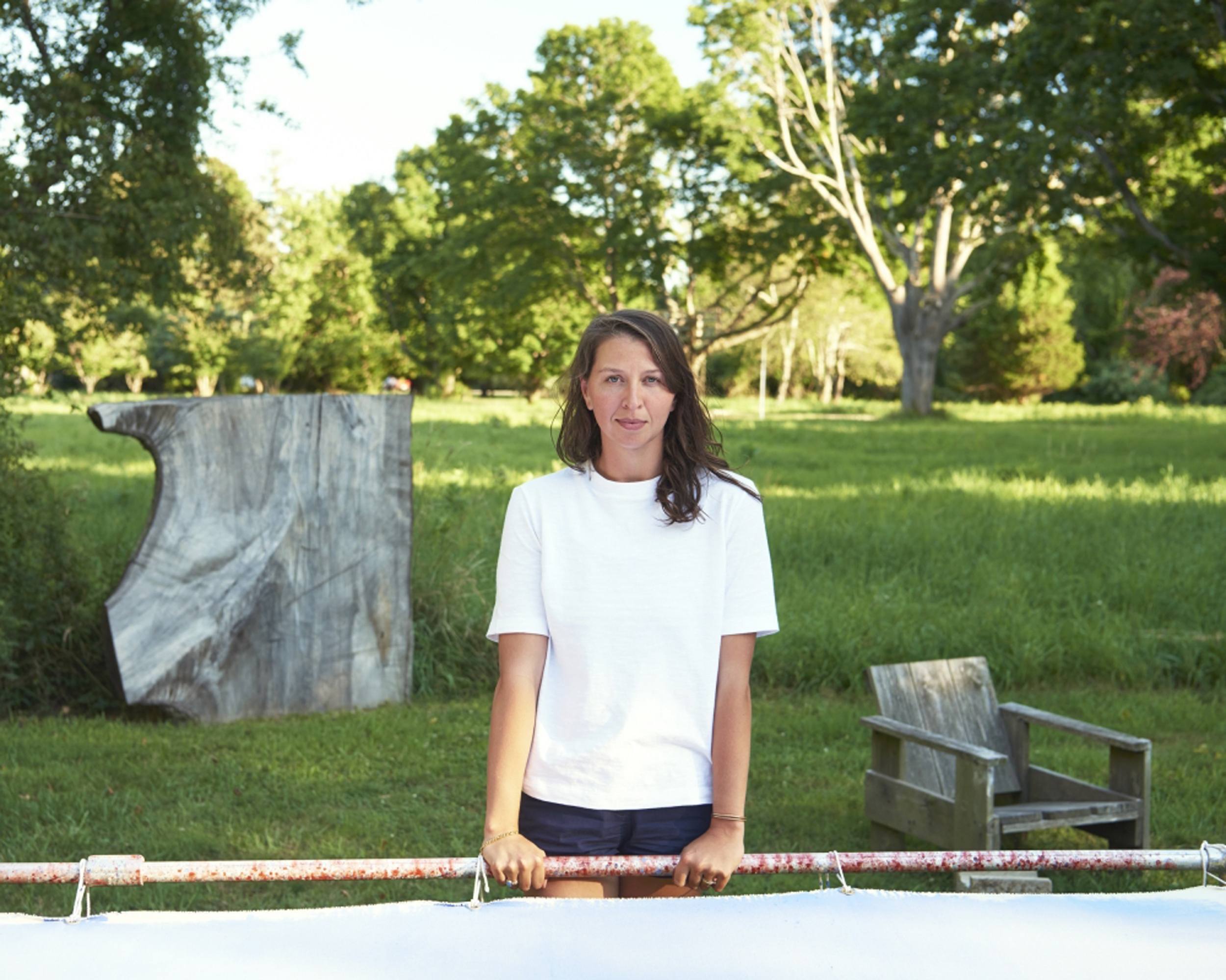 Woman in a white shirt standing outdoors near a piece of abstract art and a wooden chair, surrounded by lush greenery.