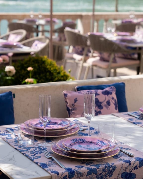 Outdoor dining set with blue and pink floral plates and glasses on a patterned tablecloth by the water.