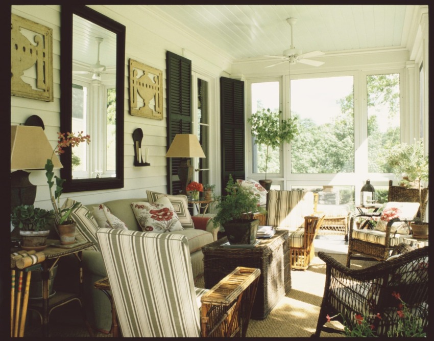 Cozy sunroom with wicker furniture, potted plants, striped cushions, and large windows overlooking a garden.