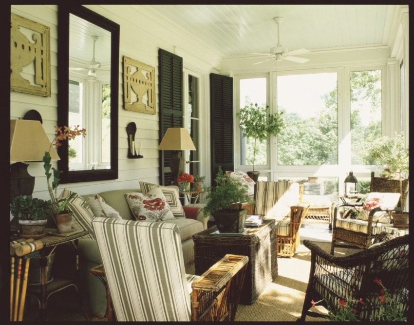 Cozy sunroom with wicker furniture, potted plants, striped cushions, and large windows overlooking a garden.