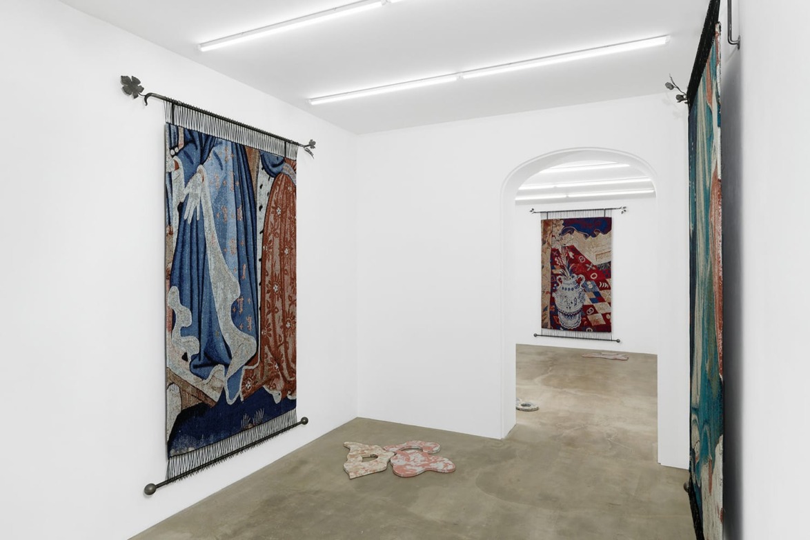Art gallery interior with large tapestries hanging on white walls and decorative pieces on the concrete floor.