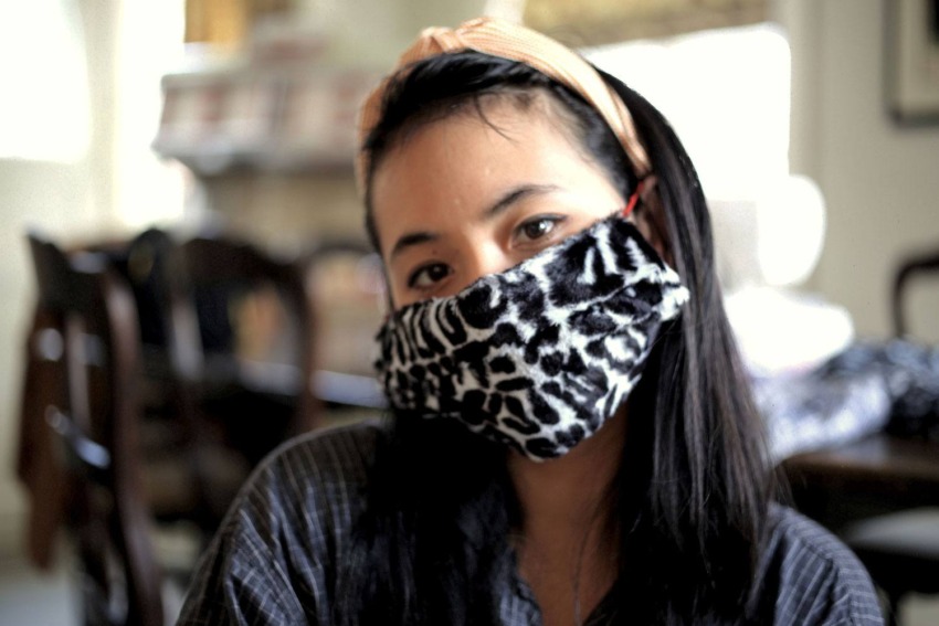 Person wearing a patterned face mask sits indoors, with a headband and long hair visible, blurred background.