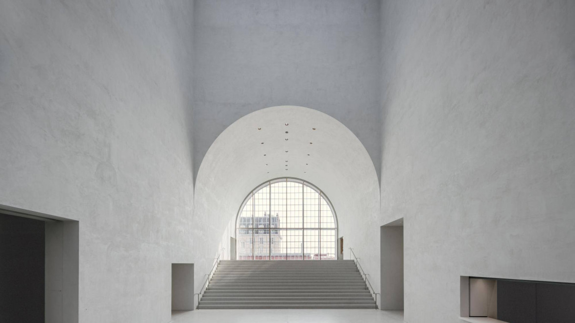 Spacious, modern interior with high arched ceiling, large window, and wide staircase leading to an upper level.