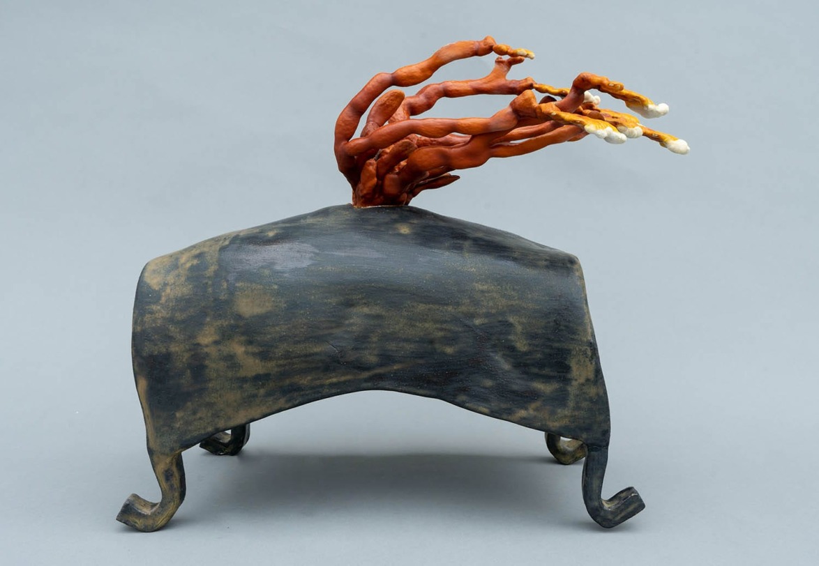 Sculptural artwork featuring a dark base with vibrant orange branching forms extending upwards against a neutral background.