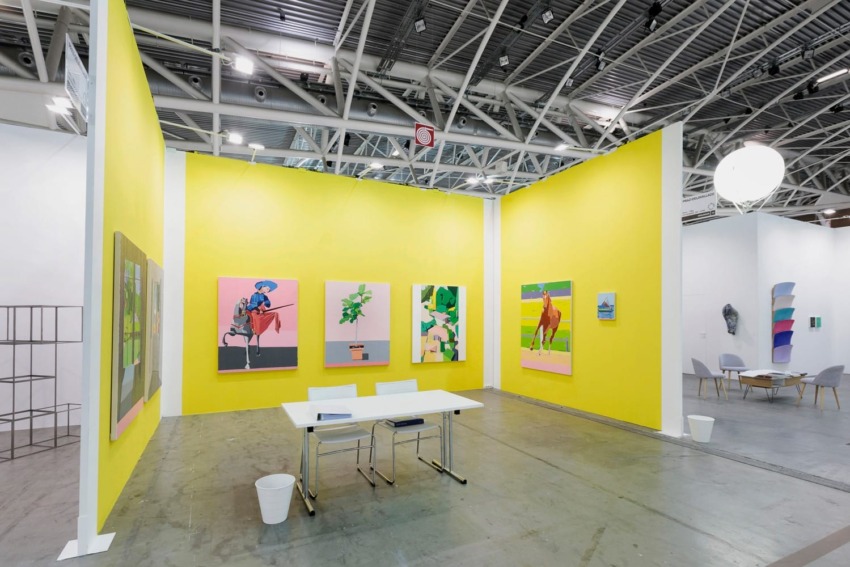 Art gallery booth with a bright yellow wall showcasing colorful paintings, white table with chairs, industrial ceiling.